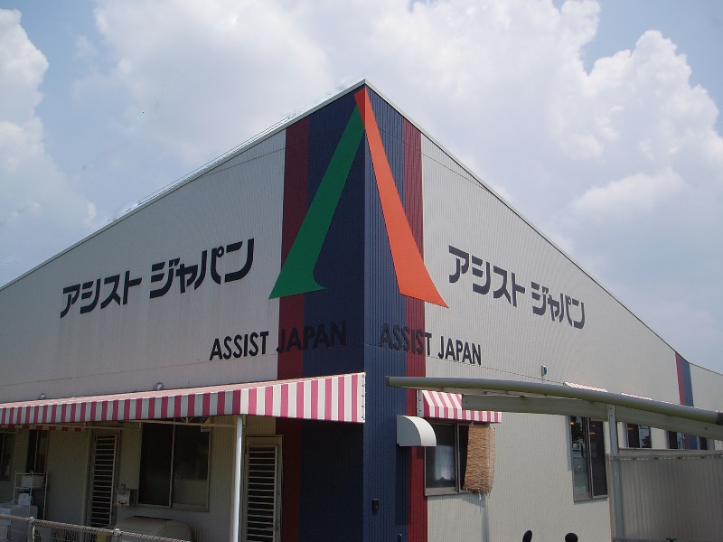 青空の下に立つアシストジャパンデイサービスセンター1号館の看板と建物全景（クリックで拡大表示）